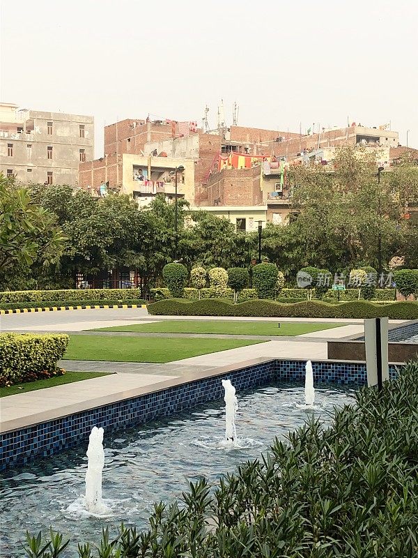 印度新德里公园的现代现代花园设计形象，有喷泉线，正式的蓝色马赛克瓷砖反射池/池塘，修剪树篱和传统的印度房屋
